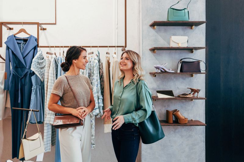 Two women engage in a pleasant conversation while shopping in a stylish boutique. They explore various fashion items together, surrounded by clothes, shoes, and bags.