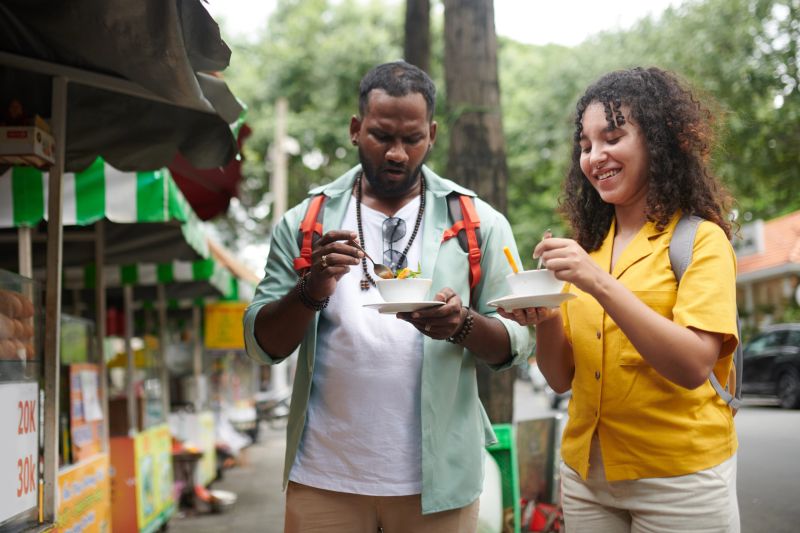 Young intercultural couple with backpacks eating street food in urban environment while standing by track during excursion