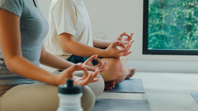 Closeup of cheerful young Asian serene couple meditates in the lotus position in a peaceful home setting, with a laptop nearby for guided meditation. Enjoying home fitness workout concept.