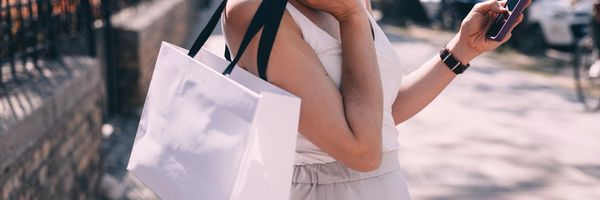 Woman carrying a white shopping bag while checking her phone outdoors.