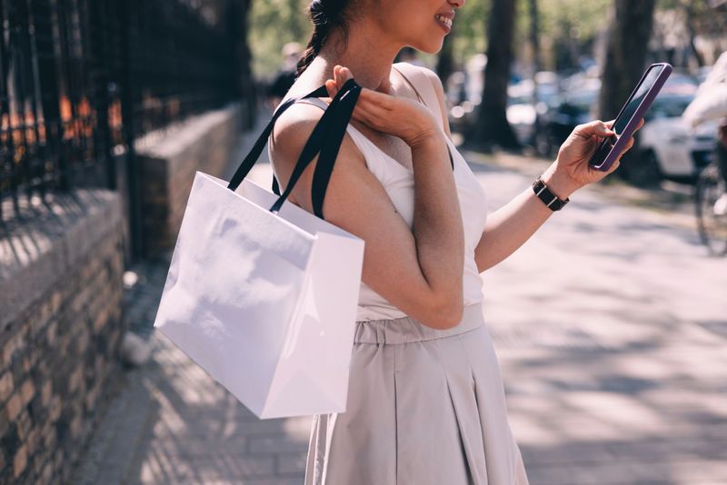 Elegant young woman in a light summer dress using her smartphone while walking with shopping bags on a sunny city street.