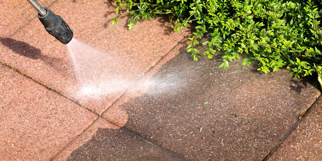 A pressure washer cleaning a brick patio near green bushes.