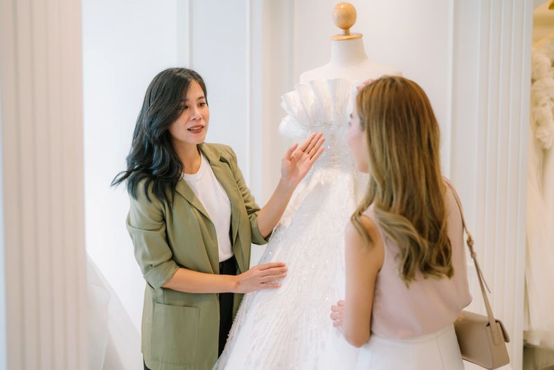 Asian bride is smiling while choosing wedding dress in modern wedding salon, Attractive designer girl using tape meter fitting on wedding dress at wedding studio