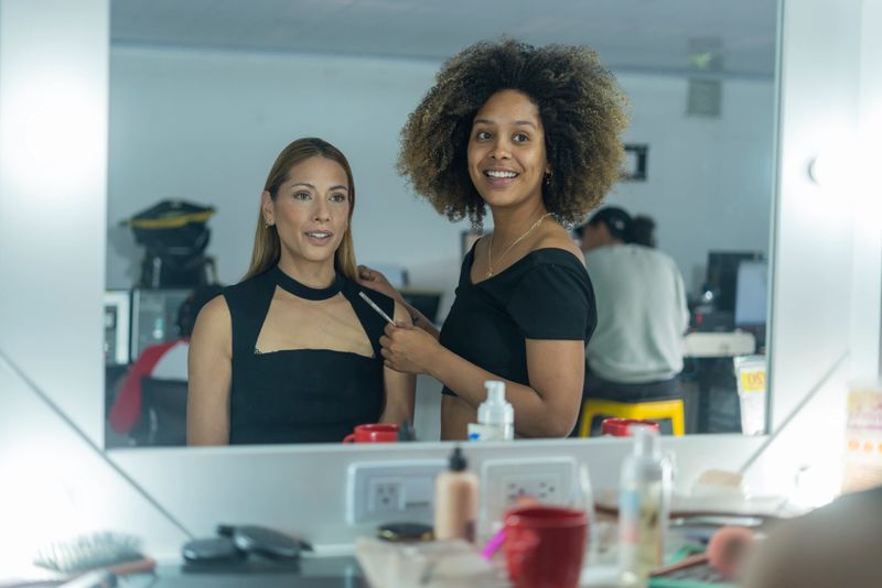 two Latin women dressed casually stand together while one of them with afro hair, a professional makeup artist, attends to her client who attends her beauty business in search of a beautiful social makeup
