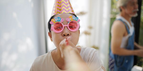 Person wearing birthday party glasses and hat blowing a party horn.