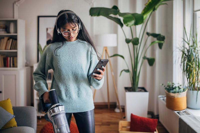 A young woman holds a cordless vacuum cleaner in one hand and a smartphone in the other in a cozy, plant-filled living room