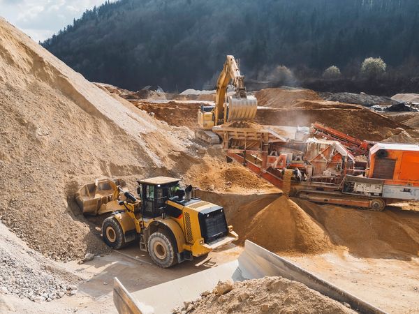 Heavy machinery working at a sandy construction site near forested hills.