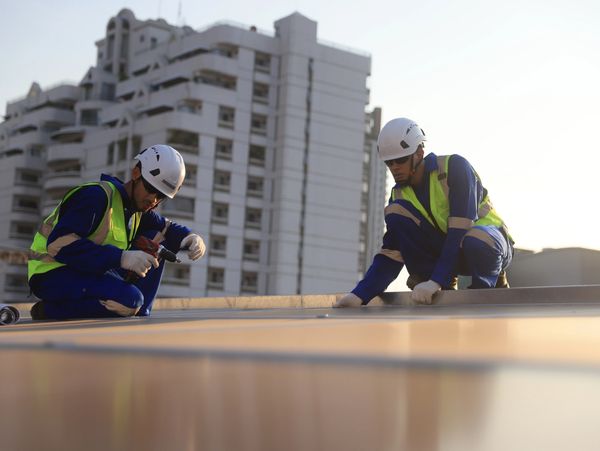 Two construction workers in safety gear working on a rooftop with a city building in the background.