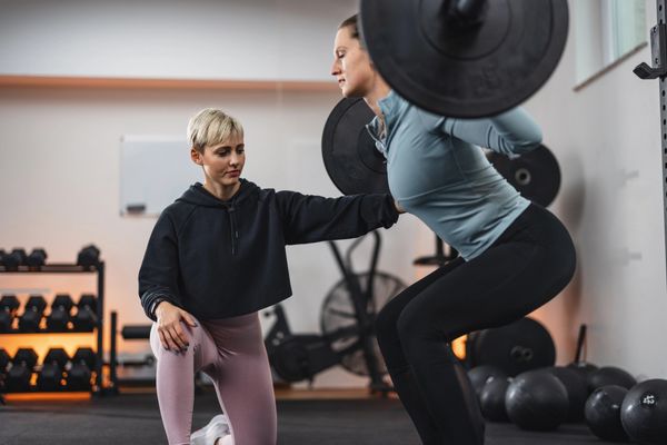 A trainer guides a woman lifting a barbell during a squat in a gym.