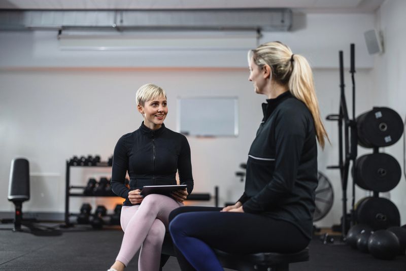 Personal trainer seated beside client in a gym, going over exercise routine and goals, in a supportive and professional environment.
