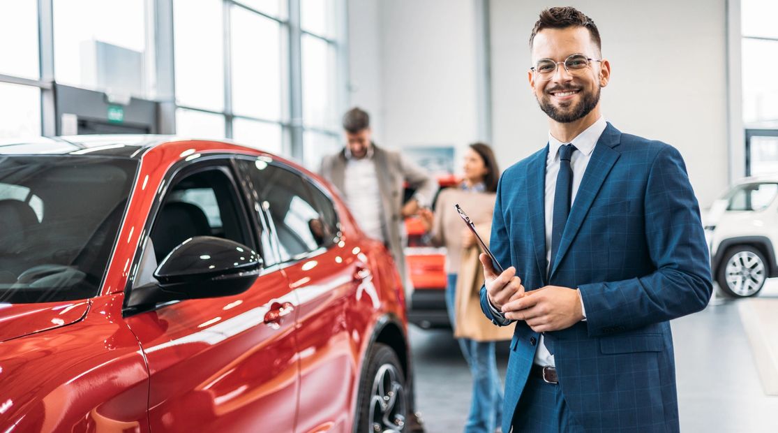 Smiling car salesman in blue suit standing by a red car in showroom.