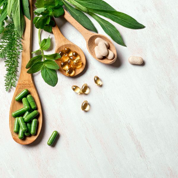 Wooden spoons with herbal capsules, soft gel capsules, and tablets alongside fresh green leaves.