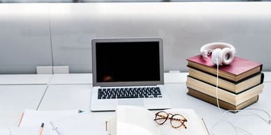 A study desk with a laptop, open book, glasses, notebooks, and headphones on stacked books.