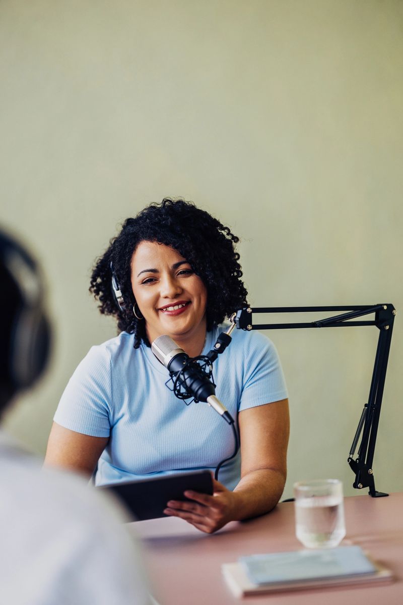 A cheerful professional woman hosts a podcast, speaking into a microphone in a studio setting. She displays confidence and engagement, making eye contact with her unseen co-host.