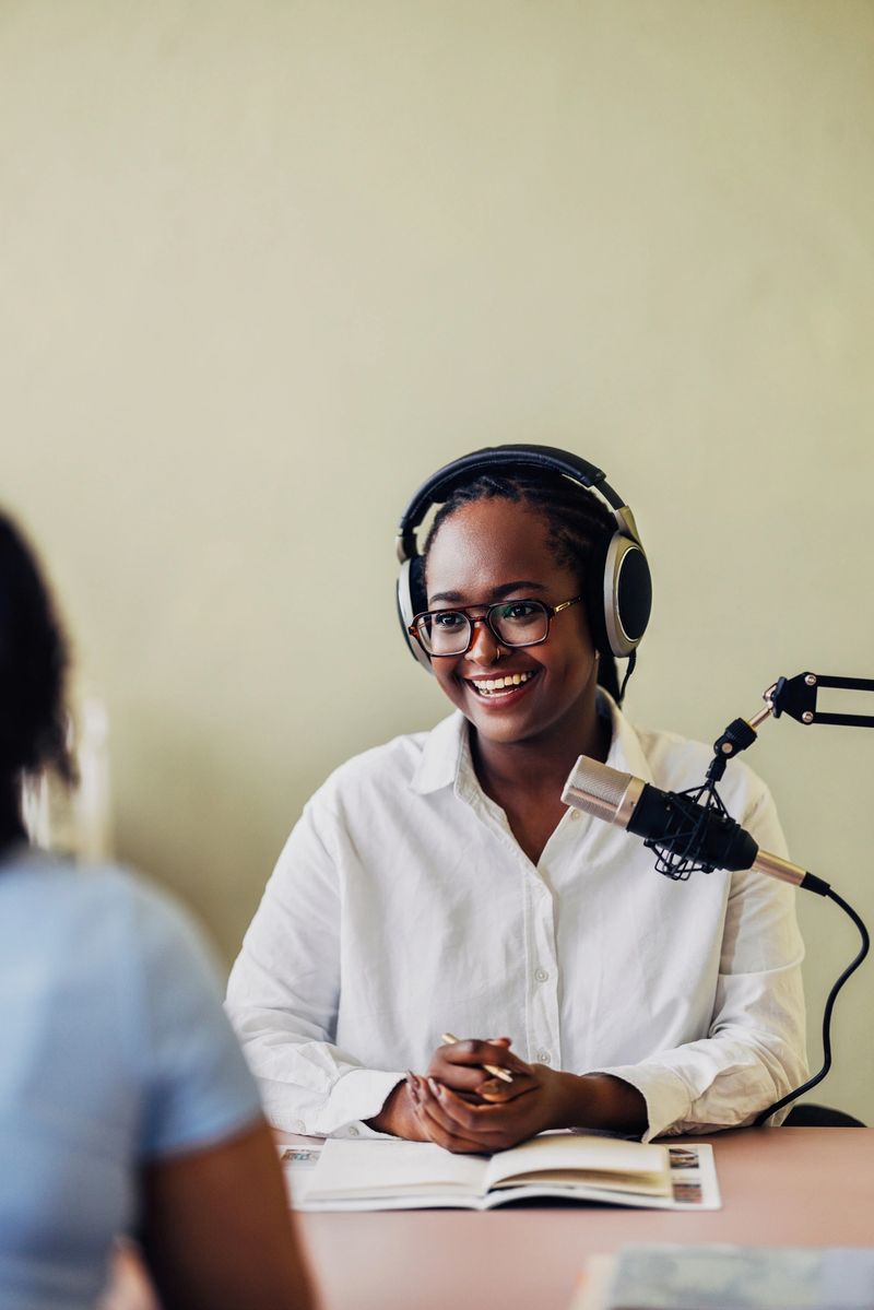 Enthusiastic young African American woman recording a podcast, wearing headphones and speaking into a microphone, engaging with a guest in a studio setting.