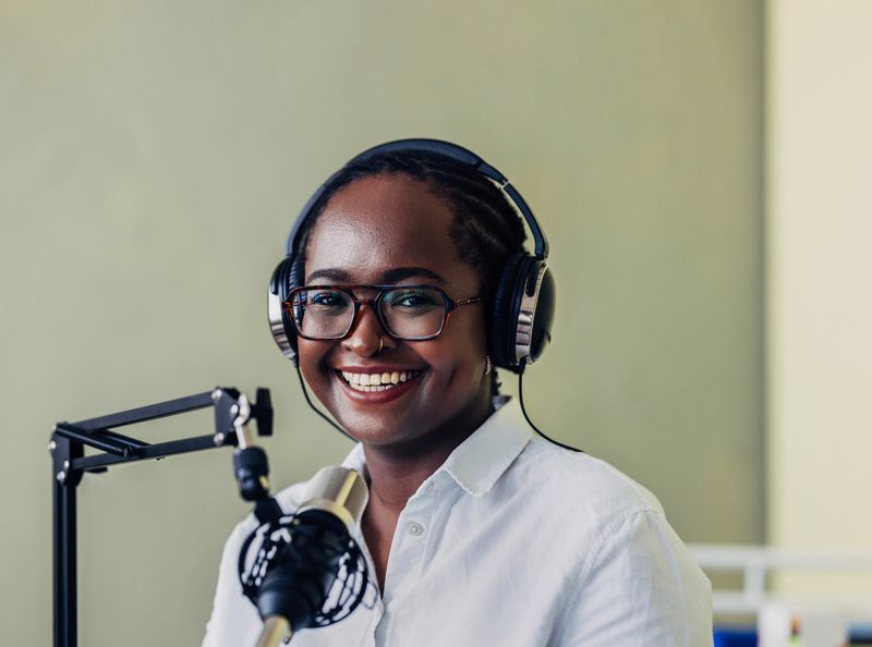 A cheerful African American woman records a podcast in a professional studio, wearing headphones and smiling, showcasing media production and communication.