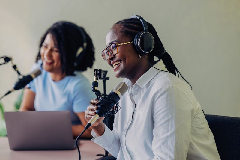 Two diverse women enjoying a lively podcast recording session. They are smiling, wearing headphones, and using microphones in a softly lit studio.