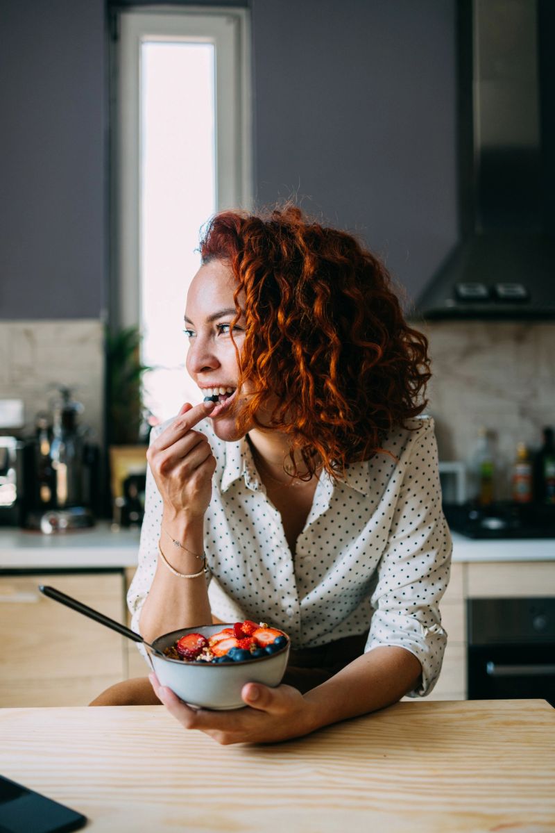A young woman with vibrant red hair is pictured enjoying a healthy bowl of fruit and yogurt in a stylish kitchen. The natural light enhances her cheerful demeanor.