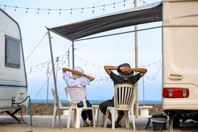 Two people relaxing on plastic chairs between RVs under string lights at sunset.