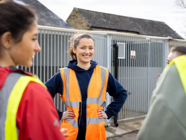A woman in an orange safety vest smiles confidently outdoors with three others nearby.