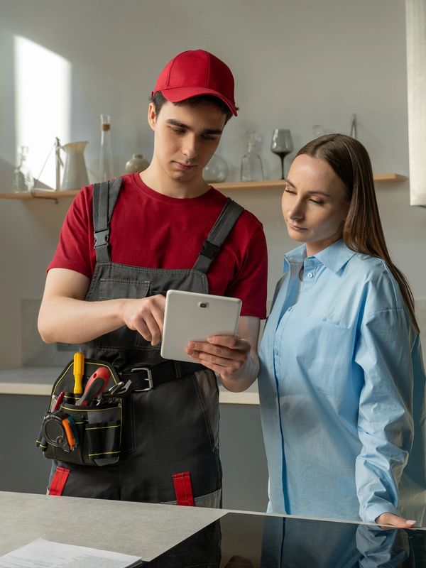 A technician in uniform shows a tablet to a woman in a modern kitchen.