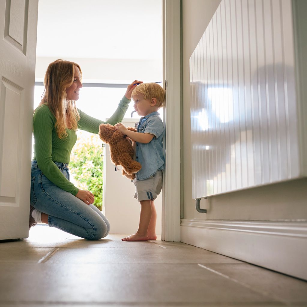 Mother measuring her toddler's height at home with a teddy bear.