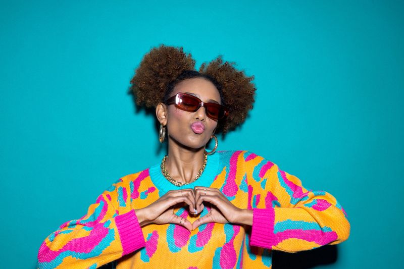 Portrait of happy gen z black young woman wearing colorful sweater and sunglasses showing heart with fingers. Studio shot, turquoise background.