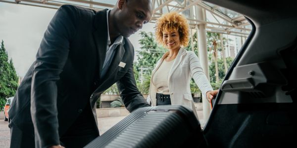 Hotel staff loading luggage into a car trunk for a smiling guest.