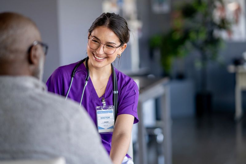 A female doctor of Hispanic decent, talks with her senior male patient during a routine check-up.  She is wearing purple scrubs and smiling as she talks with the gentleman.