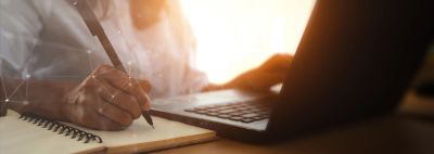 Person writing notes while using a laptop in warm light.