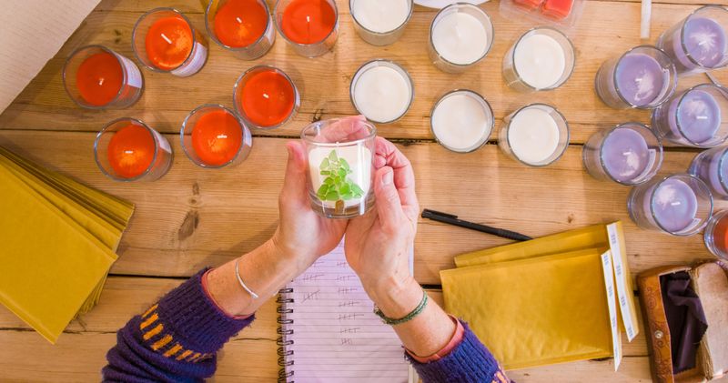 Close-up POV shot of a woman holding a candle with both hands. The candle has been decorated with colourful stones to resemble a Christmas tree. She is holding it above a wooden table that is covered with other candles and parcel packing ready to send the candles off.