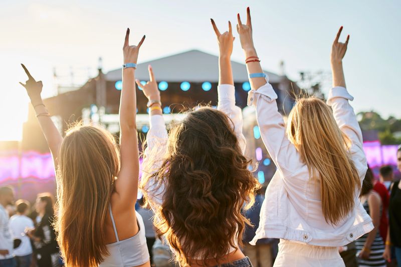 Girls dance, celebrate with hands up in summer vibe. Group of female friends enjoying live music on sunny beach festival. Casual fun, youth culture, outdoor event by sea. Friendship, lifestyle moment.