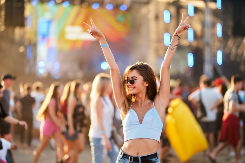 Arms raised, peace sign, dancing in crowd with stage lights. Happy young woman enjoys summer music festival on beach. Casual fashion, sunglasses, fun with friends at sunset concert party.