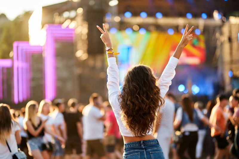 Raised hands with peace sign, enjoying live concert. Back view of young woman at summer beach music festival. Crowd, stage lights, sunset party vibes. Casual fashion, outdoor event, fun atmosphere.