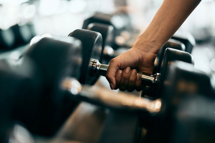 Close-up of a person’s hand gripping a metal dumbbell among a rack of weights in a gym.
