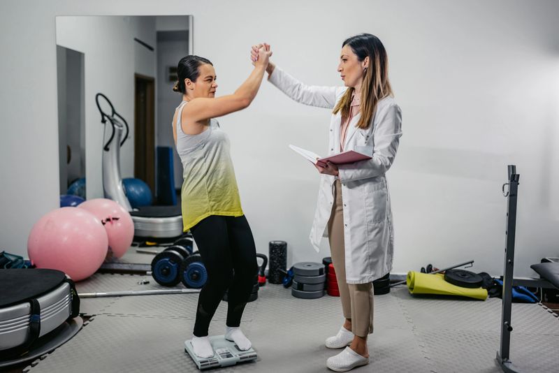 Mid-adult woman measuring her weight in the gym with her female nutritionist. Celebrating the results.