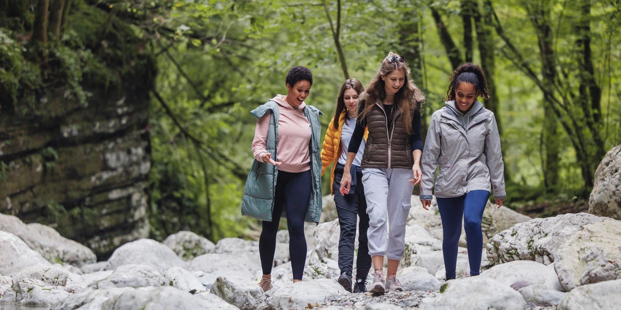 Four women enjoy a walk along a rocky riverbed in a lush forest.