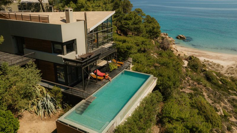 Photo of a luxury villa on a cliff that overlooks the sea. A young woman is sunbathing on the edge of the infinity pool