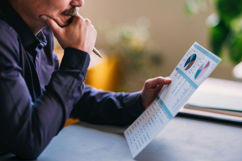 A businessman in a blue shirt thoughtfully reviews financial charts and data on a printed document, reflecting deep concentration and strategic planning in a modern office setting.