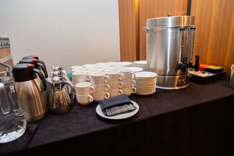 A well-organized coffee station with stacks of white cups, thermal coffee urns, and a large silver urn, suitable for events