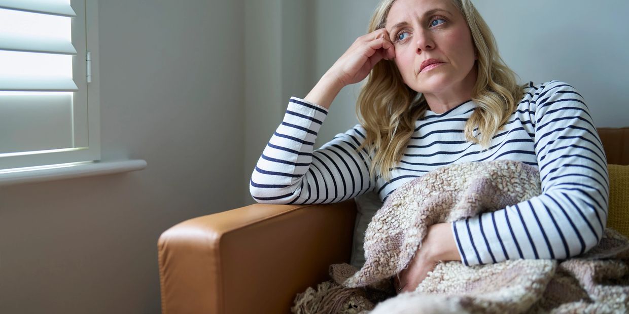 A woman looks thoughtful and concerned while sitting on a couch wrapped in a blanket.