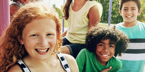 Five diverse kids smiling together at a playground slide on a sunny day.