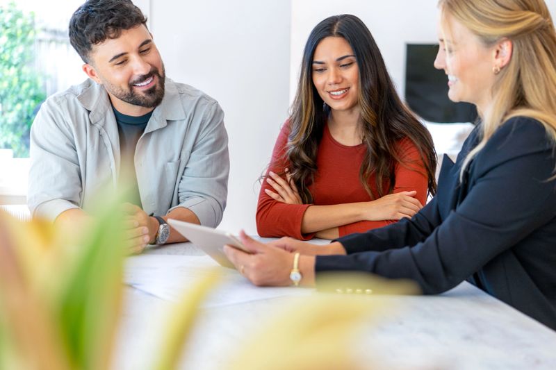 Insurance, finance or real estate agent with couple looking through documents. The agent is holding a digital tablet. Couple are casually dressed. They sitting at a table at home and are looking a little happy and smiling.