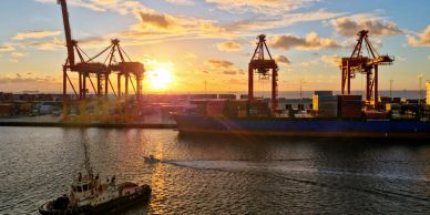 Sunset over a busy shipping port with cranes and a tugboat.