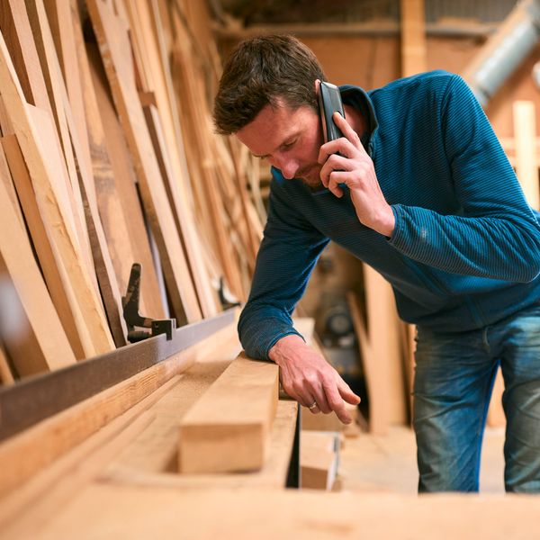 A man in a blue sweater talks on the phone while working with wood in a workshop.