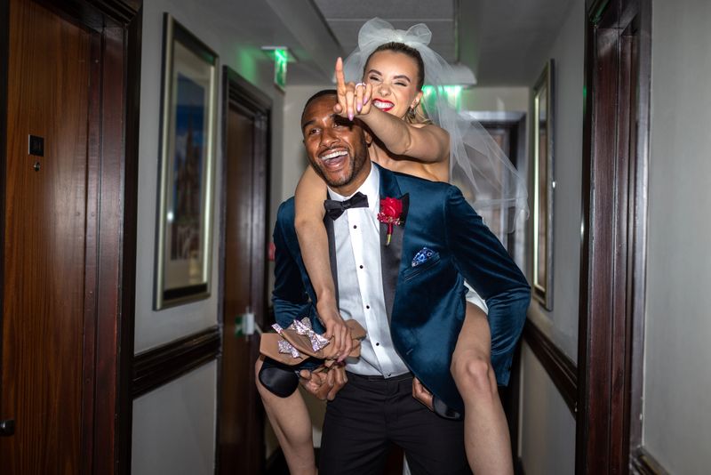 Medium shot of a newlywed couple in a hotel corridor. The young bride is piggybacking on her husband's back. She is laughing while pointing in the direction of the hotel room and carrying her high heels. The groom is carrying her while smiling.Videos are available similar to this scenario.