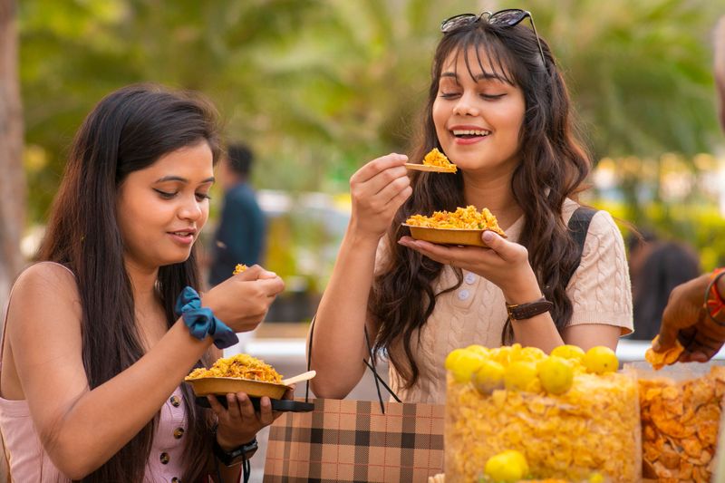 Two beautiful happy indian young women eating popular indian street snack (Bhelpuri) at street market and enjoying their weekend together at day time.