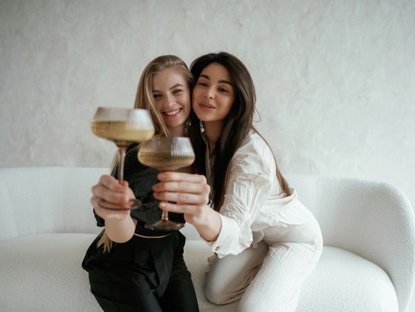 Two women smiling and toasting with champagne glasses on a white couch.