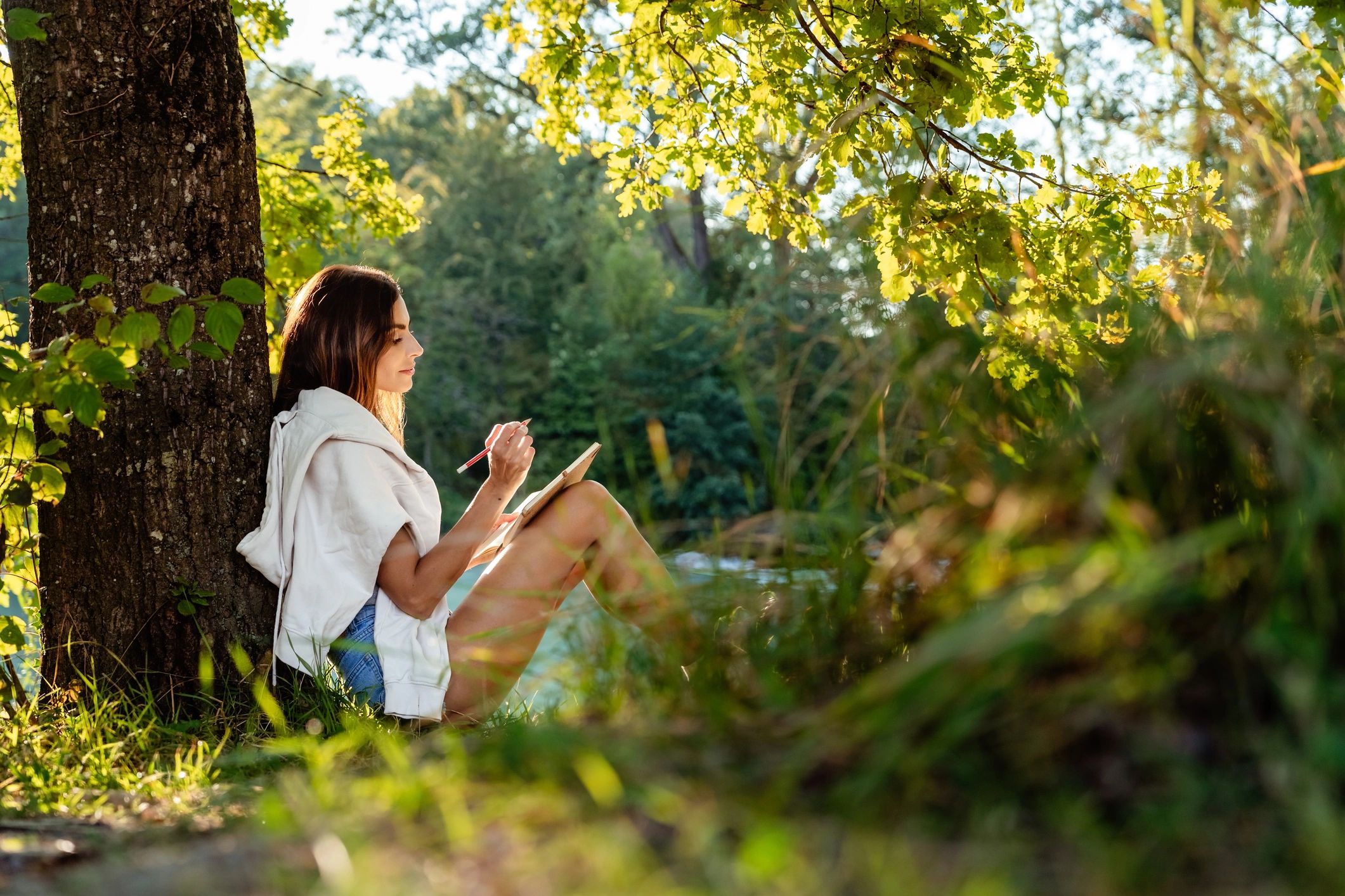 Woman sitting by a tree sketching in a notebook on a sunny day.