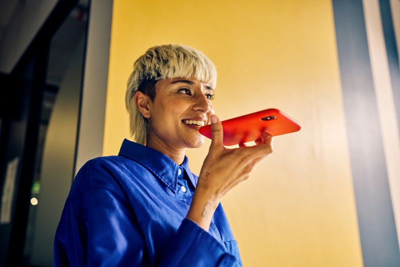 Smiling professional Asian woman using voice assistant on smartphone in a colorful office. Technology and communication in business concept